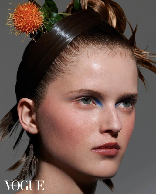 Close-up of a model with stylized hair featuring orange flower accents and bright blue eye makeup, shot in a photo studio for Vogue.