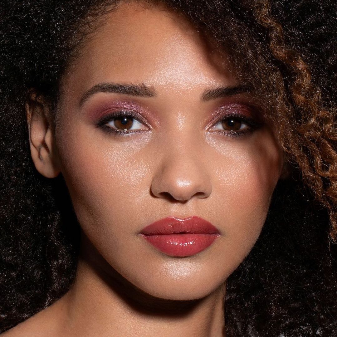 Close-up portrait of a young woman with dark curly hair, wearing pink eye shadow and lipstick against a dark background.