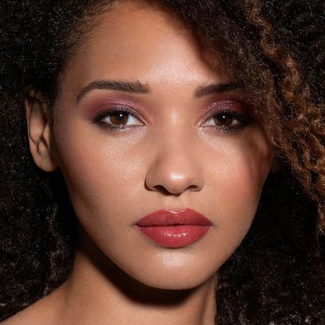 Close-up portrait of a young woman with dark curly hair, wearing pink eye shadow and lipstick against a dark background.
