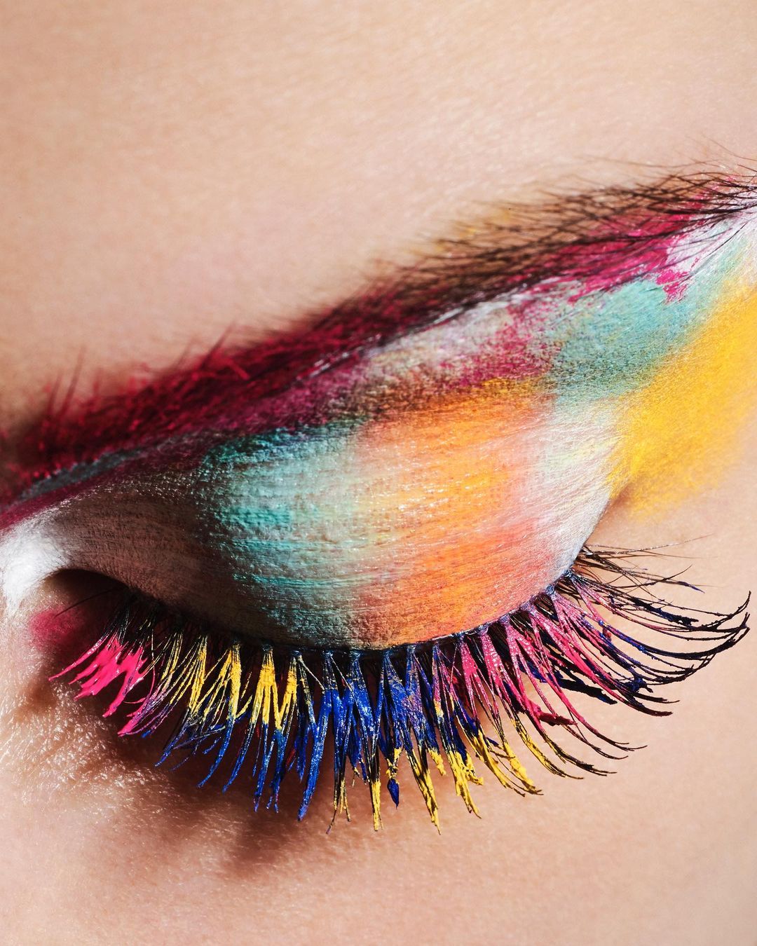 Close-up of a closed eye with vividly colored makeup applied to the eyelid and eyelashes in shades of pink, yellow, and blue.