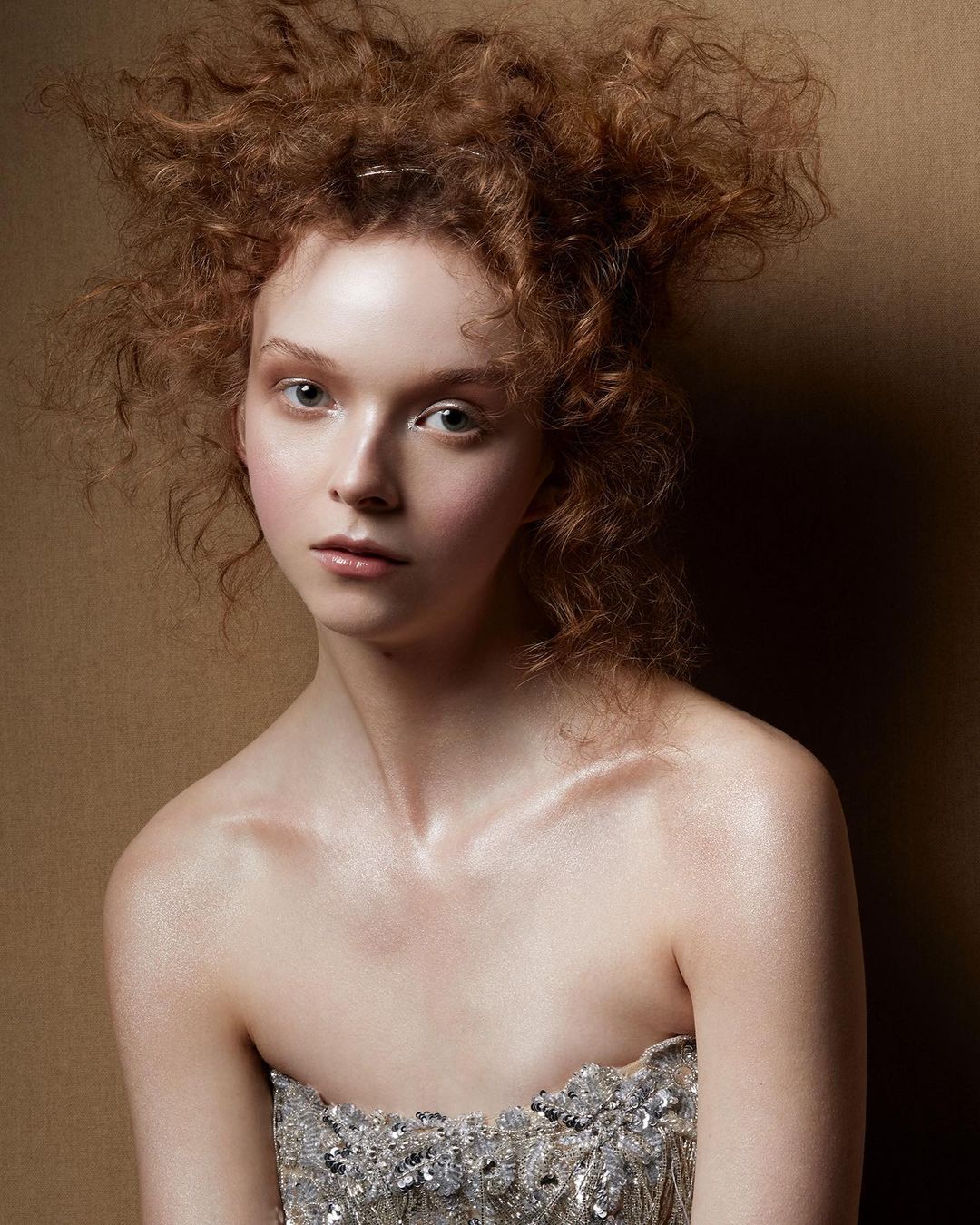 Close-up of a young adult female with reddish curly hair, wearing a silver strapless top in a studio setting with a neutral background.