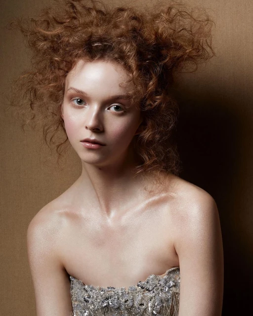 Close-up of a young adult female with reddish curly hair, wearing a silver strapless top in a studio setting with a neutral background.