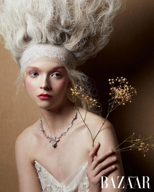 A woman with elaborate blonde hairstyle and necklace holds delicate flowers, posing against a neutral backdrop with a 'BAZAAR' logo.