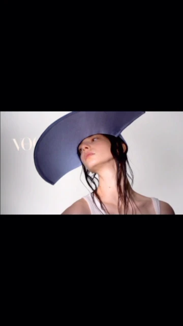 Woman with wet hair wears a large blue sun hat posing in front of the Vogue logo on a white background in a fashion shot.
