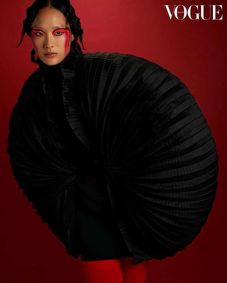 Vogue fashion editorial portrait of a woman in an oversized, pleated black jacket against a solid red background with bold red eye makeup.