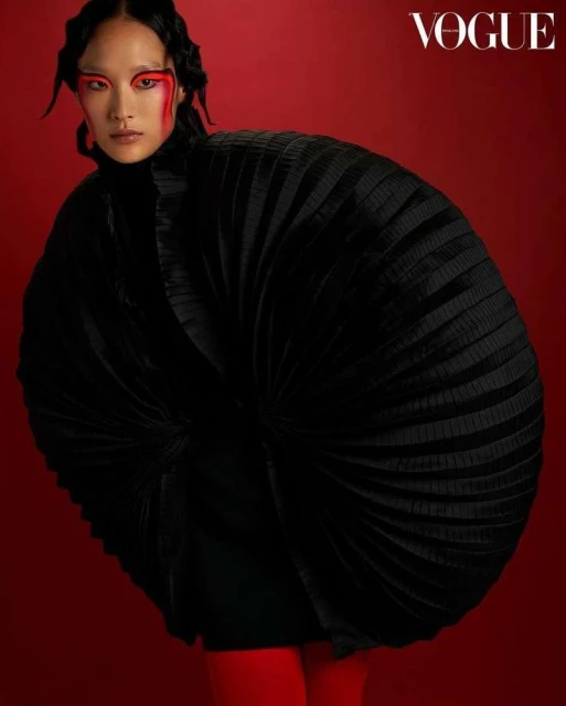 Vogue fashion editorial portrait of a woman in an oversized, pleated black jacket against a solid red background with bold red eye makeup.