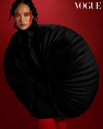Vogue fashion editorial portrait of a woman in an oversized, pleated black jacket against a solid red background with bold red eye makeup.
