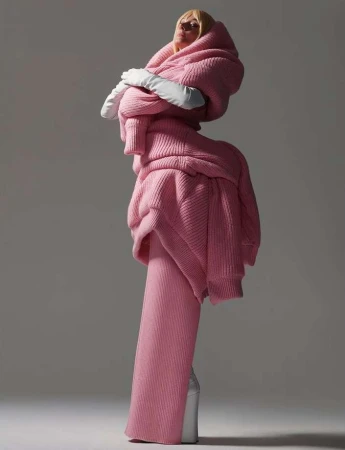 Model in voluminous pink knit sweater and matching skirt wearing white gloves and boots against a plain grey studio background