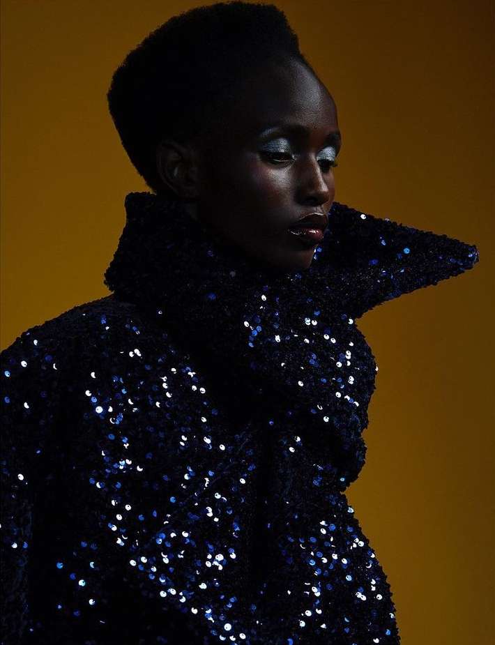 Dark-skinned woman with dramatic makeup poses in a sequined jacket against a solid yellow background in a studio shot.