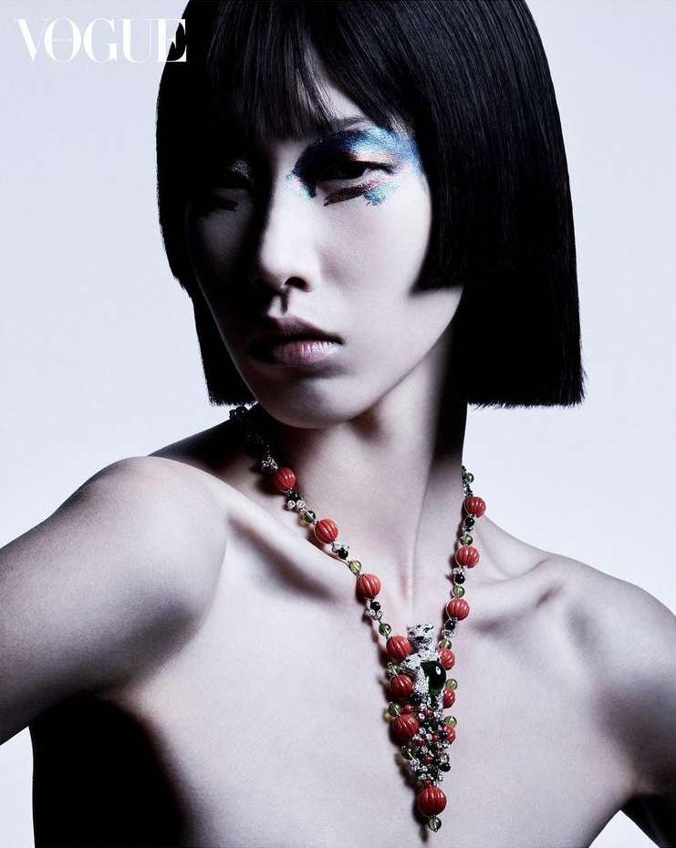 A woman with a short bob hairstyle poses wearing a beaded necklace with creative glitter eye makeup against a gradient white backdrop.