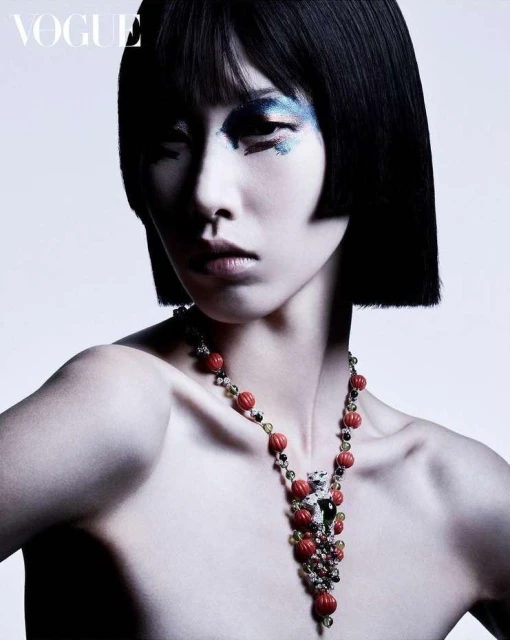 A woman with a short bob hairstyle poses wearing a beaded necklace with creative glitter eye makeup against a gradient white backdrop.