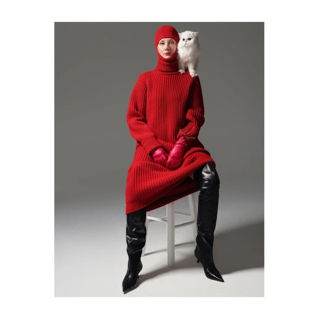 A woman in a red knit dress and hat sits on a stool with a white cat perched on her shoulder in a studio shot.