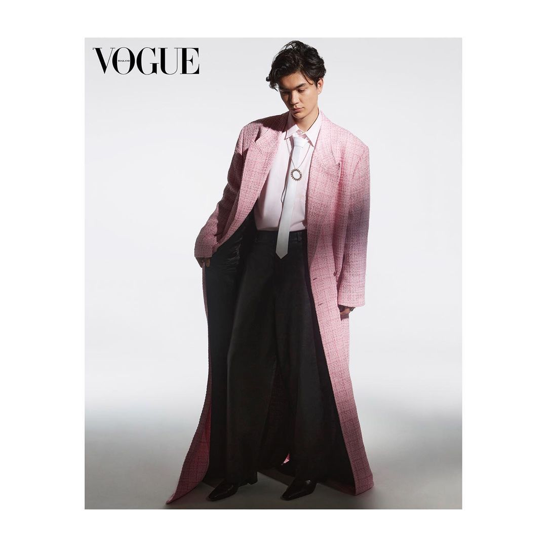 Fashion editorial shot of a young adult man wearing a pink coat, black pants, and a white tie, posing in a photo studio setting.