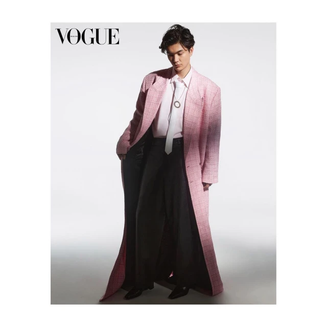 Fashion editorial shot of a young adult man wearing a pink coat, black pants, and a white tie, posing in a photo studio setting.