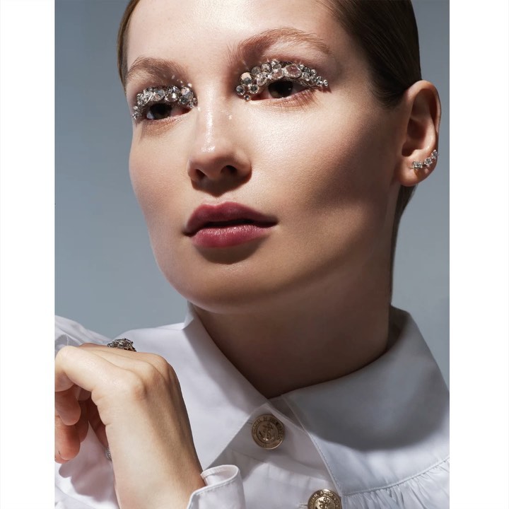 Close-up portrait of a woman wearing decorative jewels around her eyes, jewelry, and a white collared shirt against a neutral backdrop.