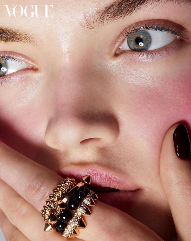 Close-up of a woman's face with dark nail polish and several rings on her finger, with the Vogue logo in the upper left corner.