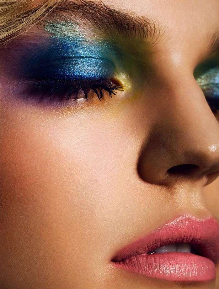 Close-up of a woman's face featuring bright blue and green eyeshadow, long lashes, and soft pink lips for a beauty campaign.
