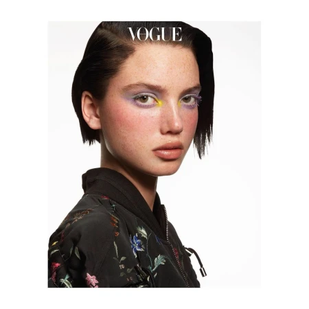Close-up portrait of a model with short dark hair, freckles, and colorful eye makeup wearing a black floral jacket against a white background.
