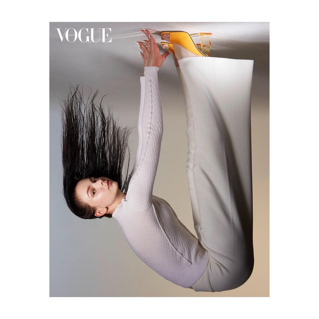 Upside-down fashion shot features a woman with dark hair, wearing white pants, a sweater, and yellow heels, on a neutral background.