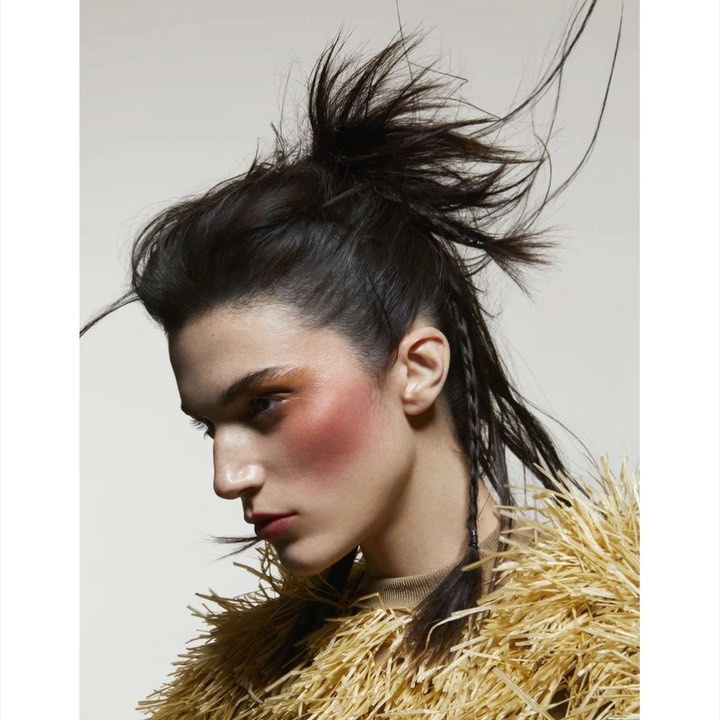 Profile shot of a woman with dark hair styled in a high, spiky topknot, and wearing a beige top with a tan textured, straw-like jacket.