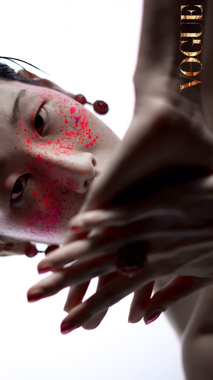 Fashion portrait: Woman with pink splattered face and painted nails posing against a white background, Vogue logo in the corner.