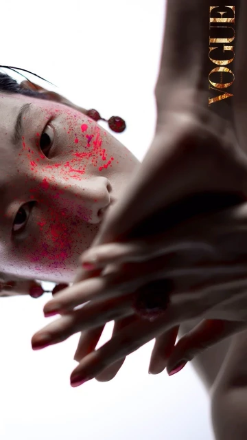 Fashion portrait: Woman with pink splattered face and painted nails posing against a white background, Vogue logo in the corner.