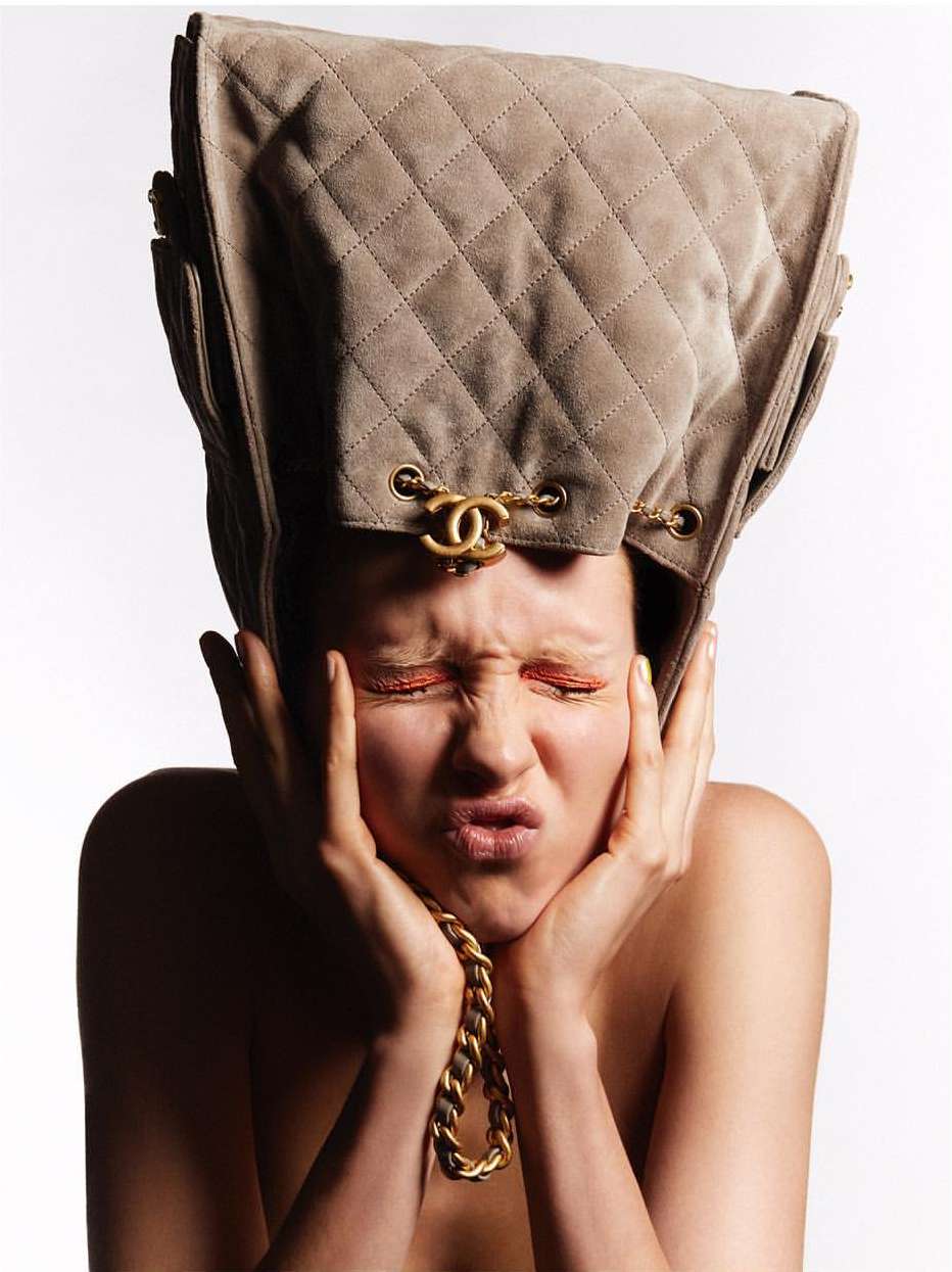 Woman with closed eyes posed with a tan quilted Chanel bag on her head and a gold chain necklace, against a plain white backdrop.
