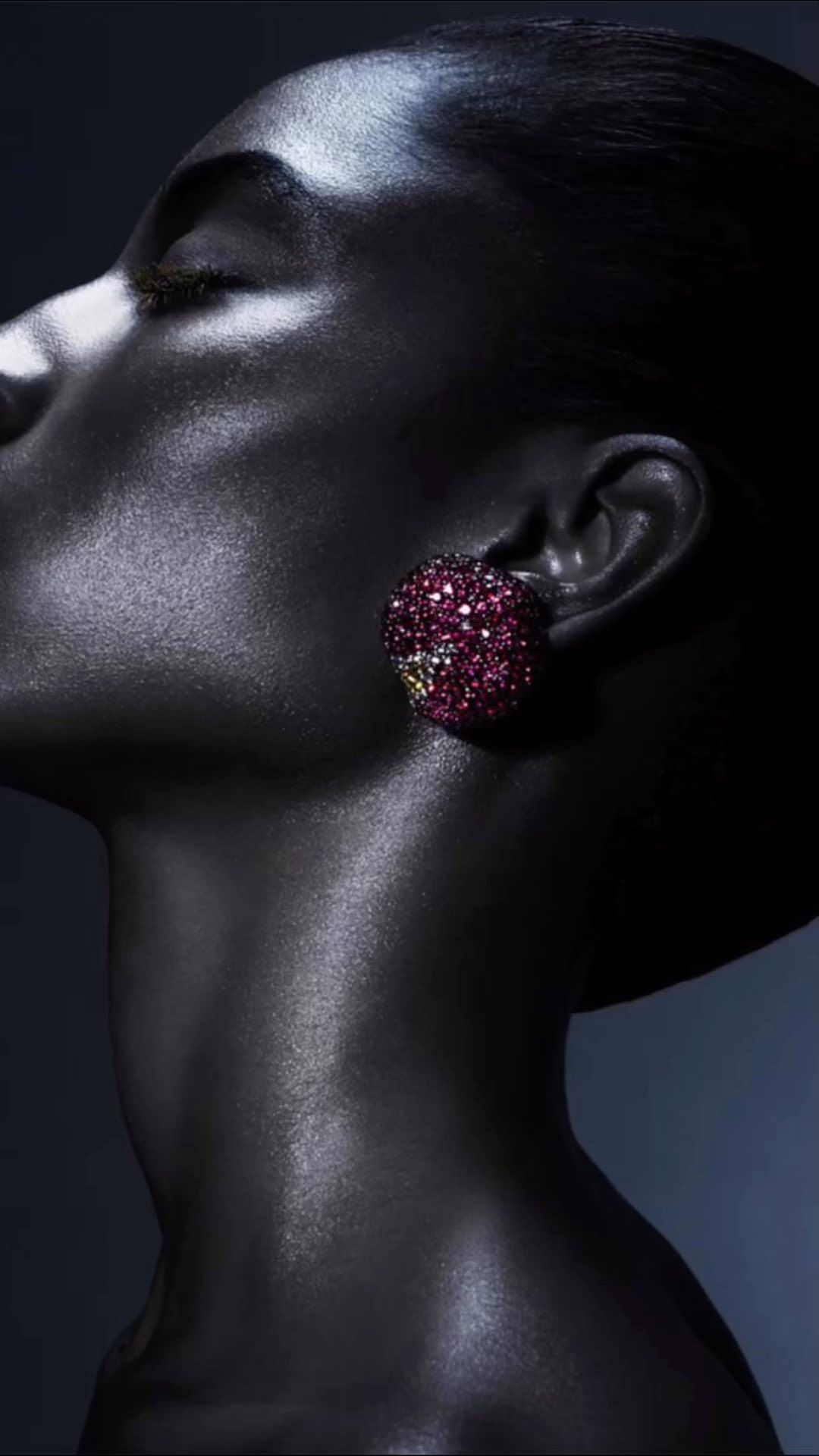 Close-up profile portrait of a black woman with glistening skin wearing a large dark pink jeweled earring by Rui Faria.