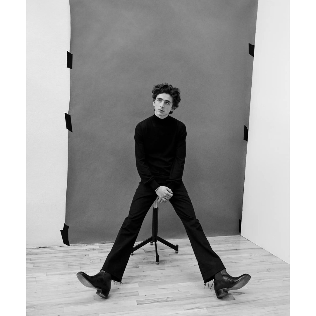 Black and white portrait of a young adult sitting on a director's chair in a photo studio against a grey backdrop.