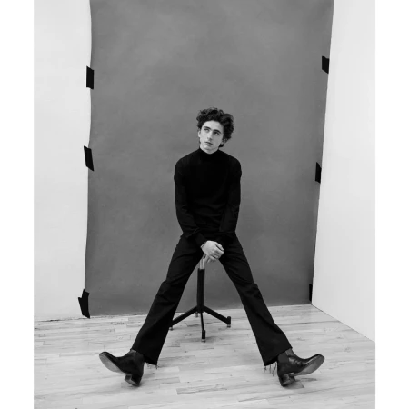 Black and white portrait of a young adult sitting on a director's chair in a photo studio against a grey backdrop.
