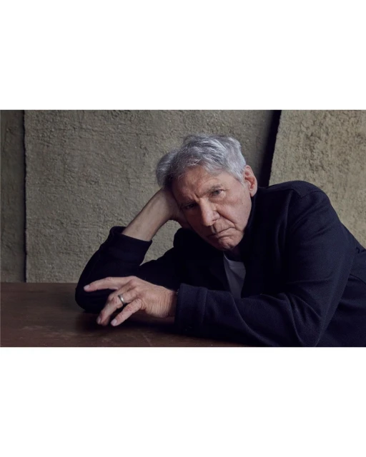 Portrait of actor Harrison Ford, grey hair, in dark jacket, leaning on a wooden table in front of a textured grey wall, looking at the camera.