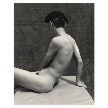 Black and white studio portrait of a nude male wearing a beret, sitting on a surface, with his back angled toward the camera.