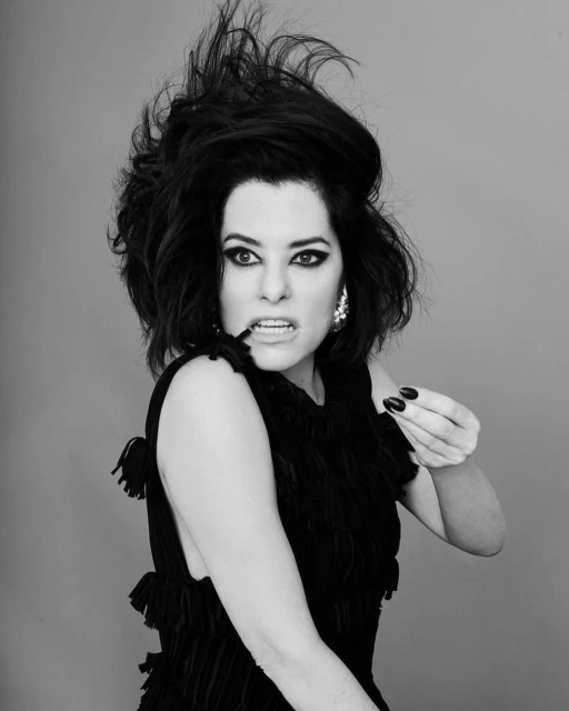 Black and white portrait of a stylish woman with dramatic hair and makeup, wearing a fringed top, against a plain gray background.