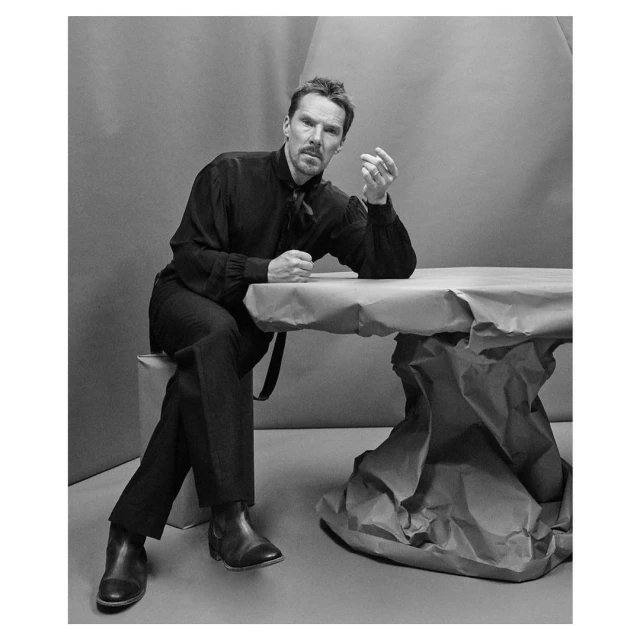 Monochrome portrait of Benedict Cumberbatch seated at a table, wearing a dark shirt and pants, set against a gray backdrop.