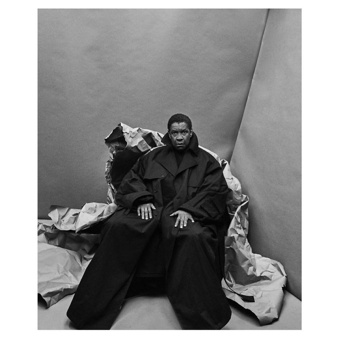Monochrome portrait of an adult with short hair wearing an oversized black coat and sitting in a gray studio with crumpled paper.