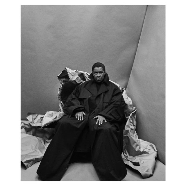 Monochrome portrait of an adult with short hair wearing an oversized black coat and sitting in a gray studio with crumpled paper.