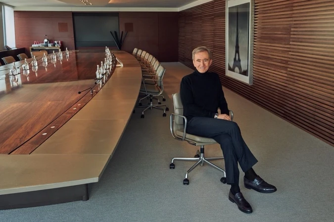 Image shows an adult male seated in a conference room in front of a long table, wearing a dark sweater and dress shoes.