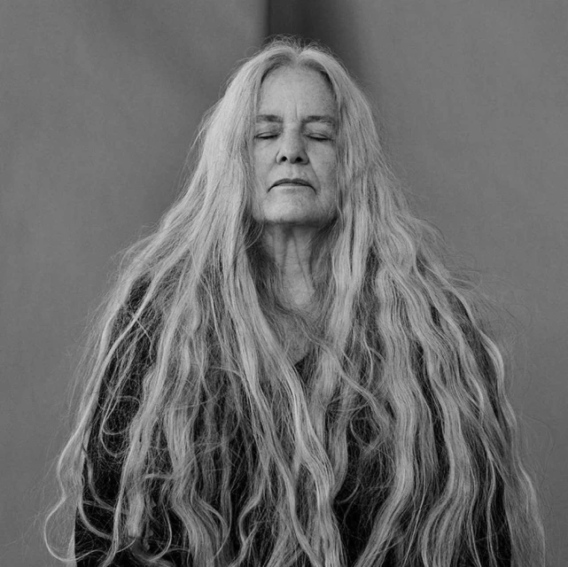 Monochrome portrait of an elderly woman with closed eyes and long, flowing gray hair against a simple, blurred studio backdrop.
