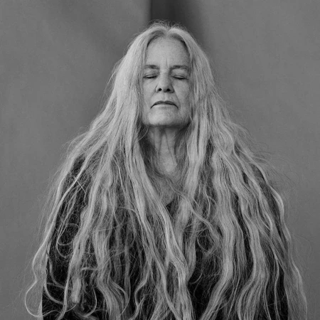 Monochrome portrait of an elderly woman with closed eyes and long, flowing gray hair against a simple, blurred studio backdrop.