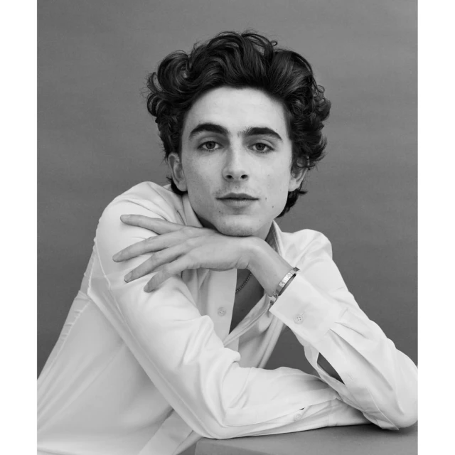 Black and white portrait of Timothée Chalamet in a white collared shirt with his hands crossed under his chin in a studio setting.
