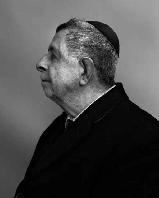 Black and white portrait of an elderly adult man in a suit coat and head covering, captured in profile view against a plain background.