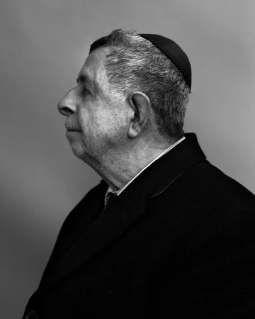 Black and white portrait of an elderly adult man in a suit coat and head covering, captured in profile view against a plain background.