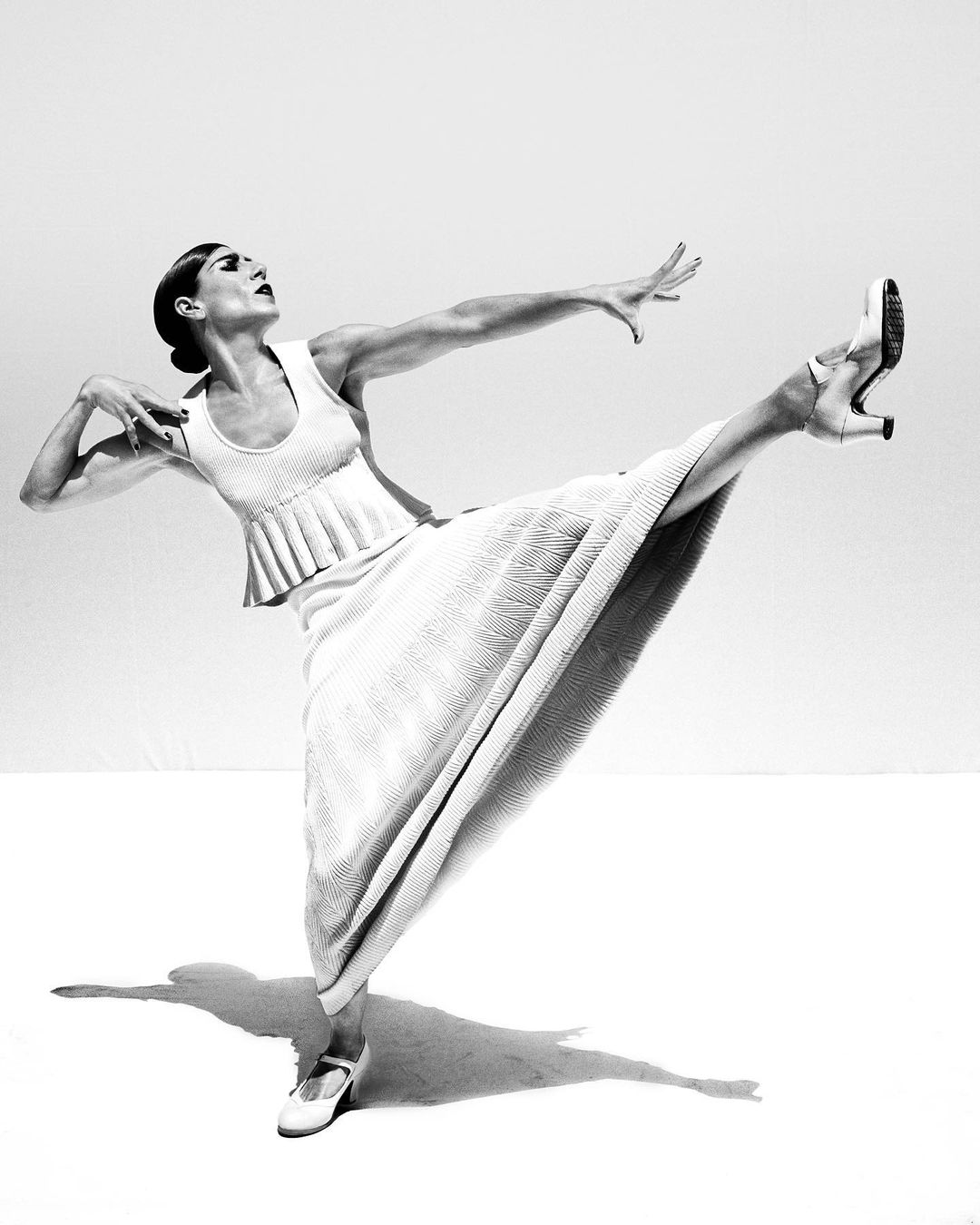 Black and white image of an adult woman with dark hair in a light-colored dress and shoes striking a pose with her leg extended.