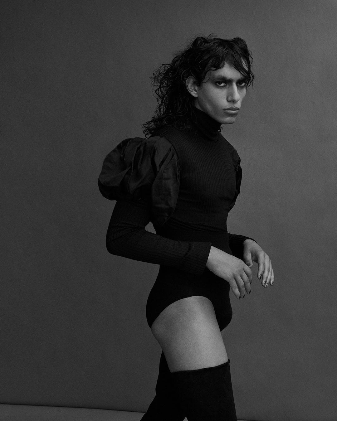Monochrome fashion portrait of a person with dark hair wearing a black bodysuit, puffy sleeves, and thigh-high boots against a grey background.