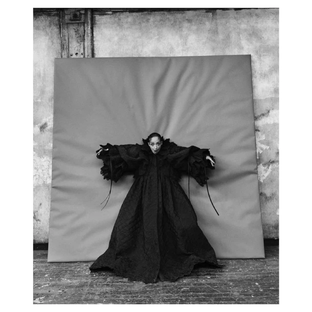 Black and white portrait of a woman wearing a dark, flowing robe against a neutral backdrop, creating a high-contrast fashion editorial image.
