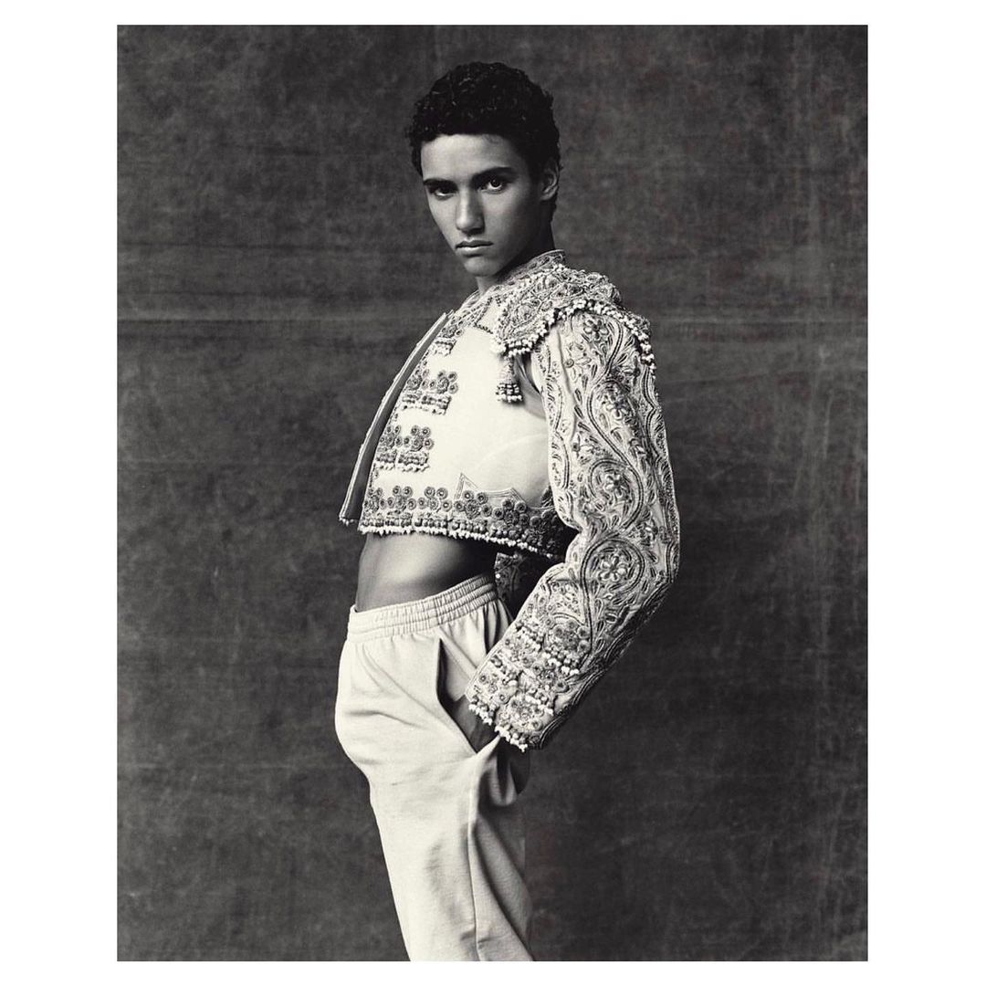 Portrait of male model in embroidered matador jacket with hand in pants pocket against dark backdrop in black and white studio photograph.