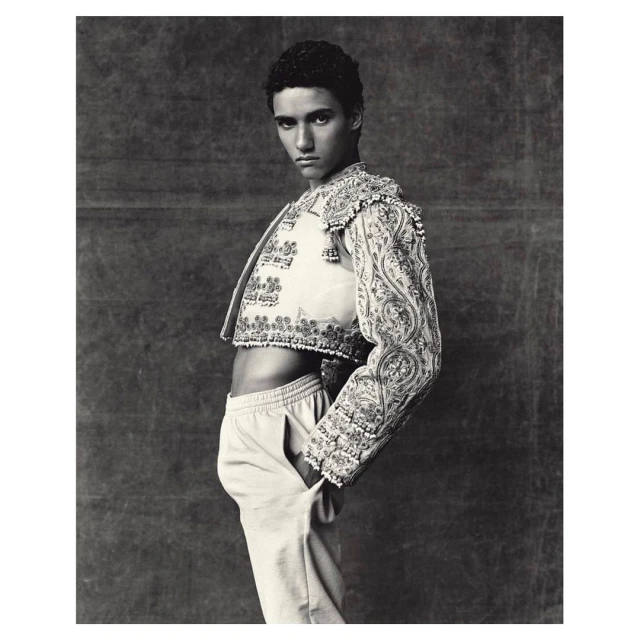 Portrait of male model in embroidered matador jacket with hand in pants pocket against dark backdrop in black and white studio photograph.