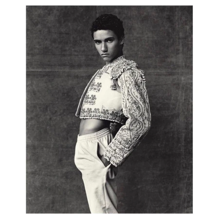 Portrait of male model in embroidered matador jacket with hand in pants pocket against dark backdrop in black and white studio photograph.