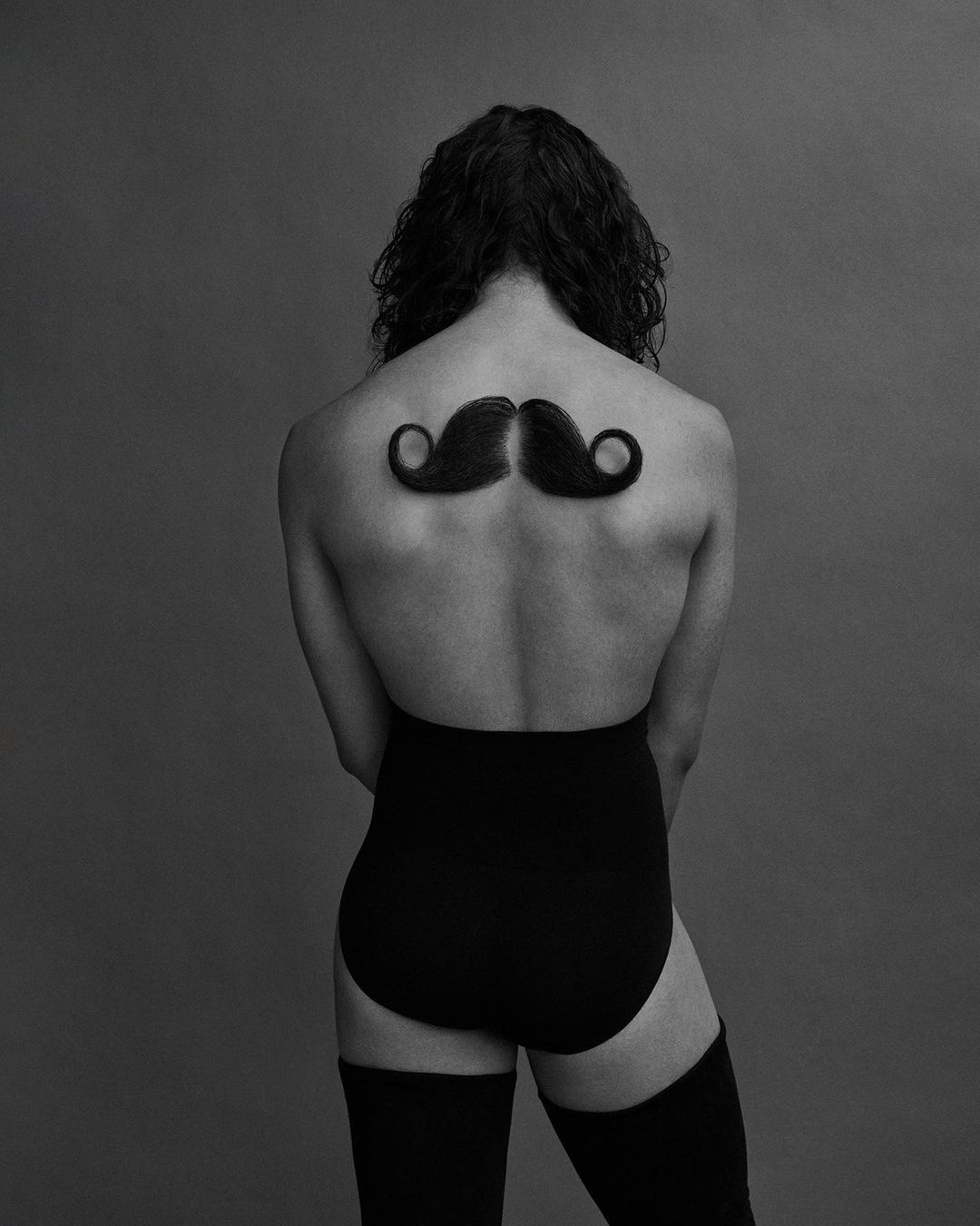 Back view of person with curly hair, wearing black bodysuit and thigh-highs, with large mustaches on their back against grey backdrop.
