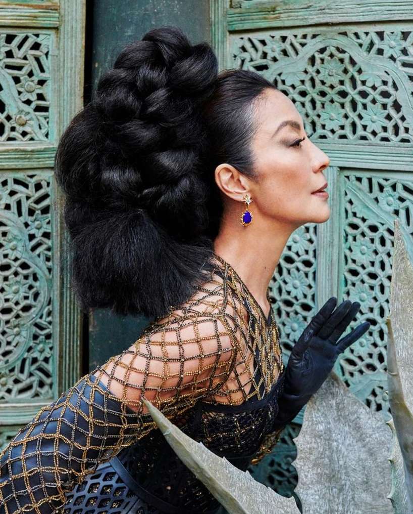 Elegant woman with a distinctive updo poses in a striking black and gold gown in front of an ornate turquoise carved door.
